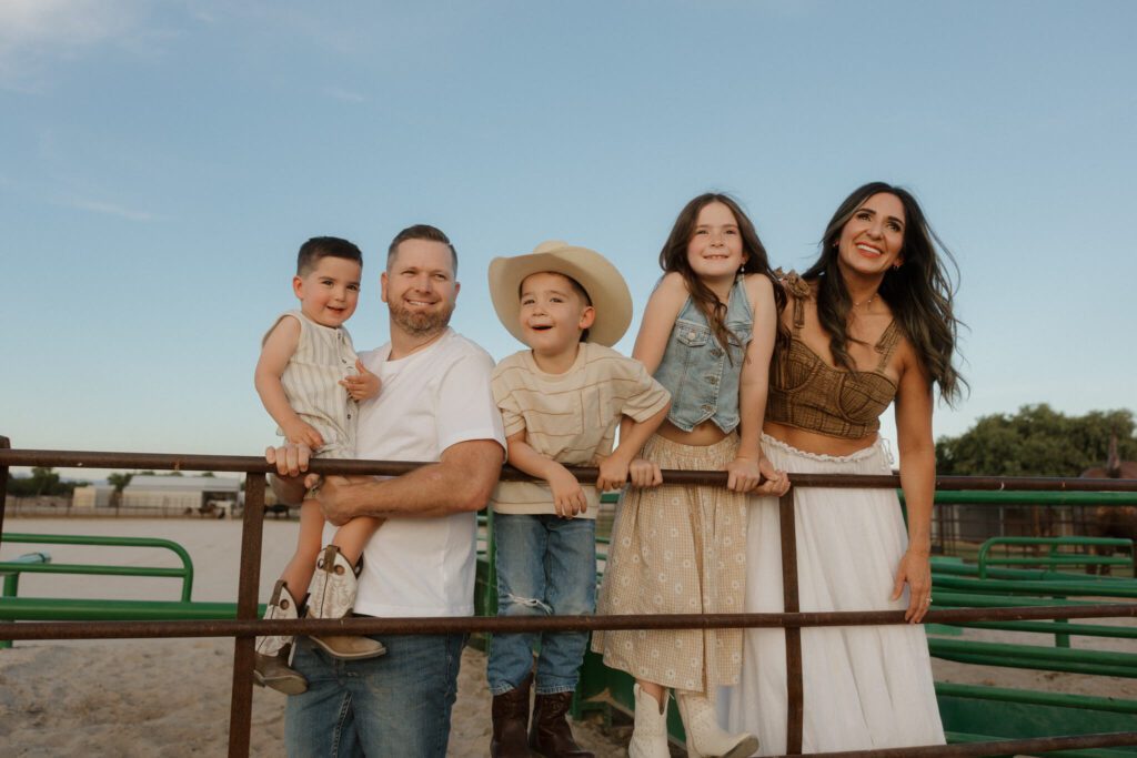 Landscape image of a family of five leaning over a farm fence smiling into the light. The family has a mom and dad with three children. They are dressed in slightly western clothing.